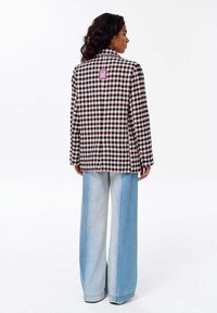 Žena s kudrnatými vlasy, oblečená v černobíle růžovém kostkovaném saku a širokých dvoubarevných světle modrých džínách, stojící zády.