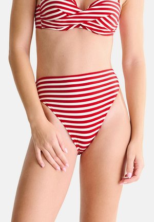 Femme portant un bikini rayé horizontalement rouge et blanc avec un bas taille haute, debout avec une main posée sur sa cuisse.