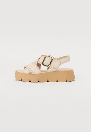 Beige platform-sandaalit ristikkäisillä remmeillä, joissa on kultainen solki ja teksturoitu ommel; paksu, uritettu kumipohja vakauden takaamiseksi.