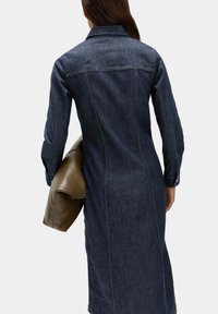 Jeansklänning i mörkblått, lång med en figursydd silhuett, knäppning vid ärmsluten, kombinerad med en ljusbrun väska. Minimalistisk design.