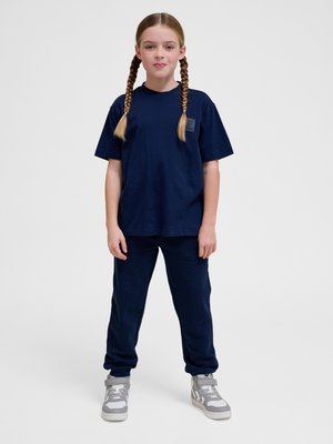 Młoda dziewczyna z warkoczami stoi, ubrana w granatowy T-shirt, pasujące spodnie i szare wysokie trampki na jednolitym tle.