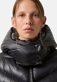 Black puffer jacket with a high, oversized collar, featuring glossy texture, prominent stitching, and large black buttons.