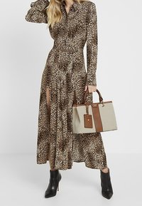 Maxi šaty s leopardím potiskem a dlouhými rukávy, s postranními rozparky, v kombinaci s béžovou a hnědou kabelkou a černými botami na vysokém podpatku.