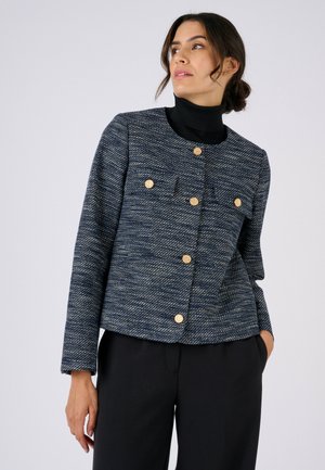 Donna che indossa una giacca blu scuro strutturata con bottoni dorati sopra un dolcevita nero e pantaloni neri, che guarda leggermente di lato.