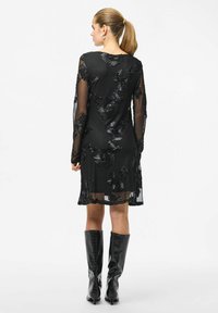 Czarna sukienka z długim rękawem, haftem kwiatowym, przezroczystą tkaniną i długością do połowy uda. Noszona z błyszczącymi czarnymi kozakami sięgającymi do kolan.