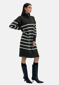 Strickkleid in Dunkelgrün mit weißen horizontalen Streifen, hohem Kragen, Viertel-Reißverschluss, geripptem Saum und langen Ärmeln. Kombiniert mit kniehohen schwarzen Stiefeln.