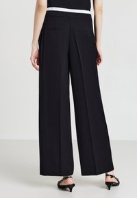 Schwarzweite Hosen mit flacher Taille, zwei Gesäßtaschen und einer glatten Textur. Kombiniert mit schwarzen Absatzsandalen.