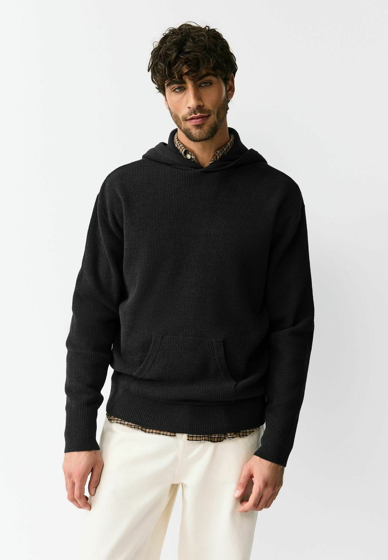 Black, textured hoodie with a front pocket and a drawstring hood, worn over a checkered shirt, paired with light-colored pants.