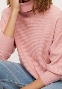 Vaaleanpunainen ribbineulottu neulepaita, jossa on korkea kaulus ja väljästi istuvat hihat, yhdistetty siniseen farkkuihin. Pehmeä tekstuuri ja rento muotoilu.