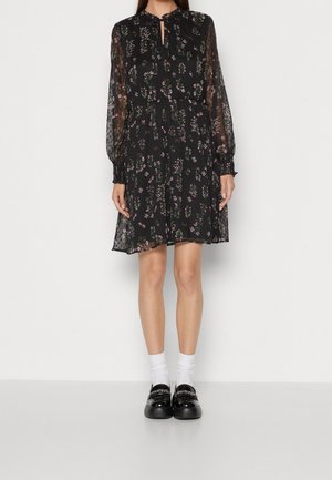 Femme portant une robe noire à fleurs mi-longue avec des manches longues transparentes, des chaussettes blanches et des mocassins noirs sur un fond blanc uni.