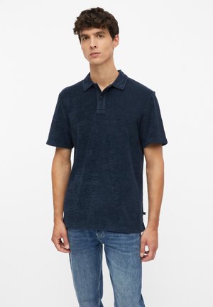 Jonge man met kort krullend haar, gekleed in een donkerblauw poloshirt met korte mouwen en lichtblauwe spijkerbroek, staand voor een effen achtergrond.