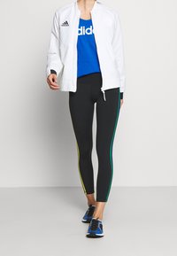 Giacca zip bianca, canotta blu con logo "adidas", leggings neri con strisce laterali gialle, verdi e blu, e scarpe da ginnastica nere.