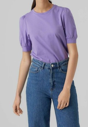 Person trägt ein lavendelfarbenes T-Shirt mit Puffärmeln, das in eine hoch taillierte blaue Jeans gesteckt ist, vor einem schlichten hellgrauen Hintergrund.