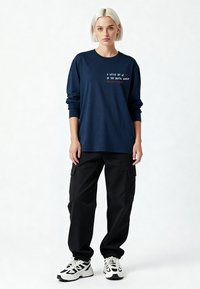Femme blonde debout portant un sweat-shirt bleu marine avec texte, un pantalon cargo noir et des baskets blanches épaisses sur un fond blanc uni.