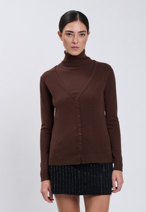 Vrouw met donker haar in een staart, gekleed in een bruine cardigan met knopen over een bijpassende coltrui en een zwarte rok met verticale witte strepen.