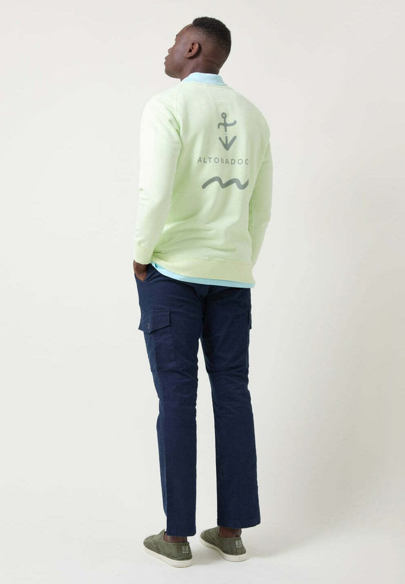 Sudadera verde claro con gráficos de ancla y ola en la parte posterior, combinada con pantalones cargo en navy y zapatillas gris. Diseño simple y ajuste casual.