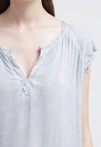 Světle modrý bavlněný top s v-neck, krajkovými detaily po obvodu výstřihu a ramenou a jemným nařasením. Krátké rukávy.