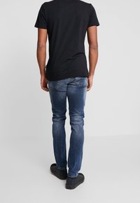 Man med svart t-shirt, blå jeans och svarta sneakers står med ryggen mot kameran framför en plain ljusgrå bakgrund.