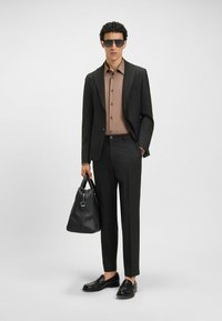 Completo nero, camicia marrone, occhiali da sole scuri e mocassini neri. Tiene una borsa nera con texture. Taglio slim, design moderno.