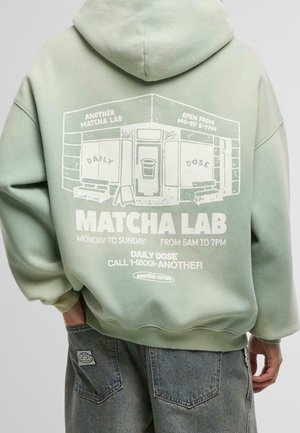 Personne portant un sweat à capuche vert clair avec un graphisme blanc d'une devanture intitulée "Matcha Lab" et les horaires d'ouverture imprimés au dos.