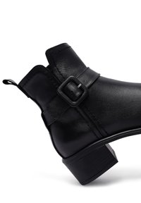 Bottine noire en cuir lisse, avec une boucle sur le côté, une tige inclinée et un petit talon empilé avec une semelle texturée.