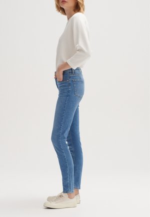 Woman wearing light blue skinny jeans, a white long-sleeve top, and beige sneakers standing sideways with hands in pockets.