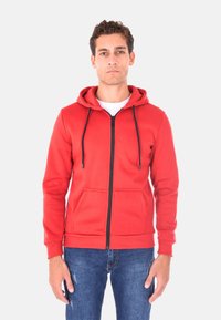 Felpa rossa con zip e cappuccio regolabile, dotata di due tasche frontali e una cerniera nera. Texture liscia; indossata sopra una maglietta bianca e jeans blu.