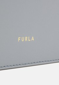 Pelle grigio chiaro a pebbles con logo "FURLA" dorato. Presenta cuciture visibili e una texture liscia, evidenziando un design minimalista.