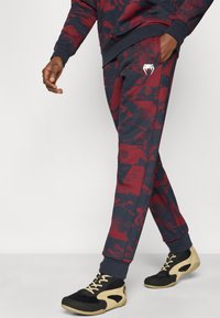 Κόκκινα και ναυτικά παντελόνια jogger με μανσέτες στους αστραγάλους και πλαϊνή τσέπη με λευκό λογότυπο. Συνδυασμένα με μαύρα και ταμπά αθλητικά παπούτσια.