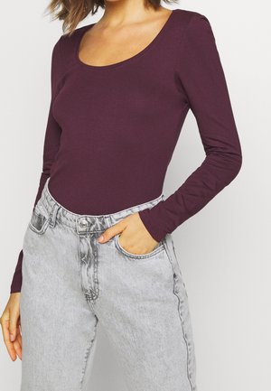 Kobieta ubrana w dopasowaną, długorękawową, ciemnofioletową bluzkę wpuszczoną w jasnoszare dżinsy z wysokim stanem, z jedną ręką w kieszeni, kadrowana powyżej ramion.