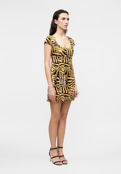 Γυναίκα που φοράει κοντό, εφαρμοστό φόρεμα με κίτρινες, μαύρες και μπεζ αφηρημένες ρίγες, συνδυασμένο με μαύρα σανδάλια με λουράκια και ψηλό τακούνι.