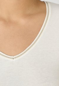 Top de cuello en V de color crema claro con una textura suave, que presenta un elegante bordado en plata a lo largo del escote. Sin patrones ni gráficos visibles.