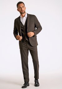 Bruin pak met een gestructureerde blazer en een bijpassend vest, gedragen over een wit shirt; gecombineerd met zwarte dresschoenen en een moderne pasvorm.