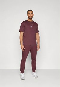 Bordó sportos öltözék, amely egy rövid ujjú pólót tartalmaz egy kis fehér logóval és hozzáillő, szűkülő szárú nadrágot, puha anyagból készült.