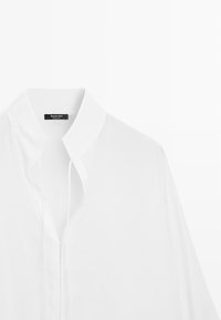 Chemise blanche à col boutonnée en tissu lisse avec une étiquette noire à l'intérieur du col indiquant le nom de la marque.