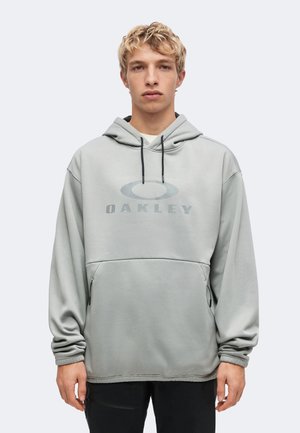 Ung mand med kort blondt hår iført en lysegrå Oakley hoodie med sorte snore og sorte bukser, stående mod en ensfarvet baggrund.
