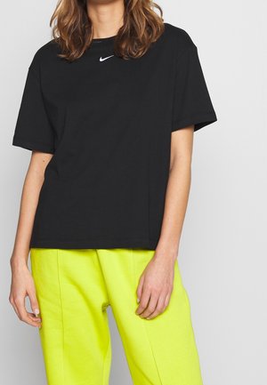 Persoon die een zwart Nike T-shirt en felgele broek draagt, staand tegen een effen lichtgrijze achtergrond.