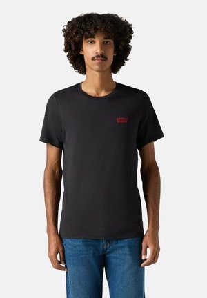 Mand med krøllet hår iført en sort Levi's t-shirt og blå jeans, stående mod en ensfarvet hvid baggrund.