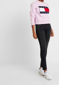 Sudadera de un rosa claro con un logotipo de marca en azul marino, combinada con jeans ajustados negros y zapatillas blancas. Atuendo simple y casual.