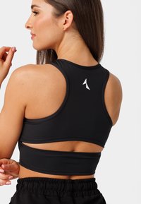 Melns sporta krūšturis ar elegantu dizainu un izgrieztu muguru, izgatavots no gludas auduma un akcentēts ar baltu logotipu.