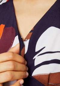 Blumenmusterstoff mit tiefblauem Hintergrund, der rote, braune und weiße Designs zeigt. V-Ausschnitt mit dekorativen Strassdetails.