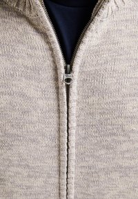 Svetle šedý pletený svetr na zip s texturovaným vzorem a kontrastní tmavou vnitřní vrstvou viditelnou na límci. Stříbrné zipové kování.