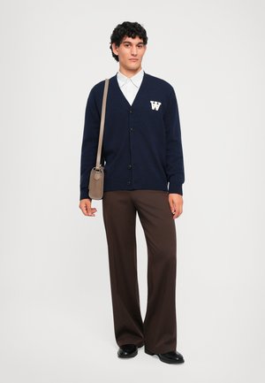 Marineblauer Cardigan mit weißem "W"-Logo, Knopfverschluss und gerippten Bündchen, kombiniert mit braunen weiten Hosen und schwarzen Schuhen. Die Accessoires umfassen eine kleine beigefarbene Tasche.