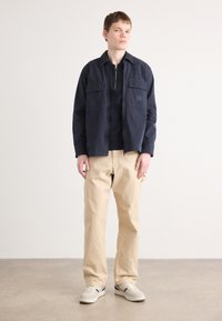Marineblauwe jas met twee borstzakken, gecombineerd met lichtbeige broek en off-white sneakers met donkere accenten. Eenvoudig, gestructureerd ontwerp.