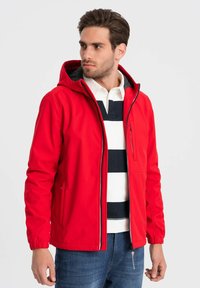 Rød softshell-jakke med hætte, lynlåslukning foran og sidelommer, parret med en mørkstribet polo t-shirt og blå jeans.