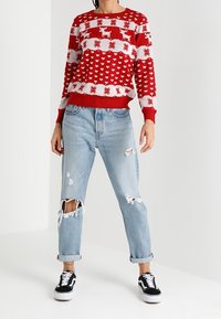 Pull rouge et blanc à motifs de rennes et de cœurs, associé à un jean bleu clair déchiré et à des baskets noires.