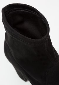 Bottine en daim noir avec un talon bloc, des coutures visibles et une ouverture supérieure arrondie sur fond blanc.