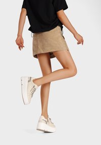 Mini gonna in suede beige con spacco laterale abbinata a sneakers di colore chiaro con dettagli metallici e suola bianca spessa.