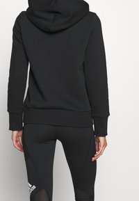 Černá mikina s texturovaným povrchem, vybavená kapucí a žebrovanými manžetami. Nosená s odpovídajícími černými legínami, které ukazují štíhlý střih.