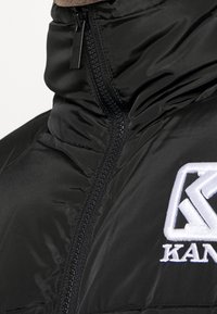 Giacca a vento nera con zip parzialmente chiusa, logo ricamato bianco sul petto che recita "KANG."